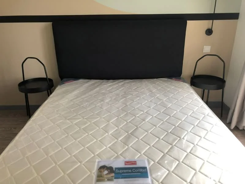 Bedroom features: king-size bed, upholstered headboard in black, two black bedside tables, lamps on bedside tables, white mattress with quilted cover, wooden flooring, decorative wall with abstract design, curtains, wall-mounted light fixtures; style: modern.