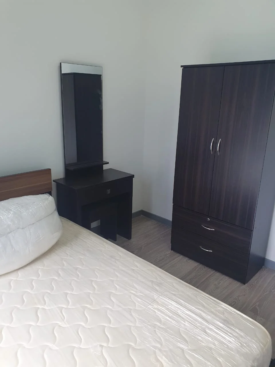 Bedroom features: queen-size bed with mattress, wooden headboard, dark wood wardrobe with two doors, dark wood dressing table, mirror on dressing table, laminate flooring, light grey painted walls, window with natural light; style: modern.