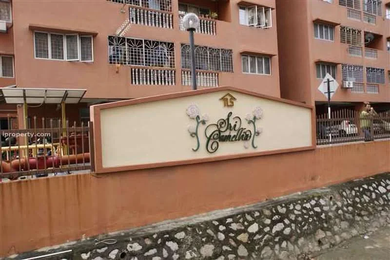 Exterior type: low-rise apartment block; features: decorative sign with 'Sri Camellia', brick facade, multiple windows with grilles, covered parking area, surrounding fence, street signage, landscaped area with stones; style: modern.