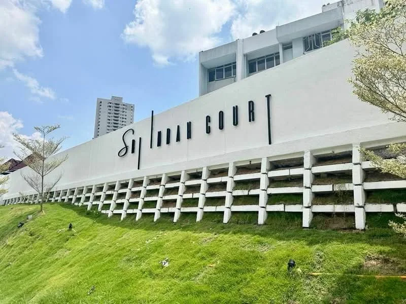 Exterior type: low-rise apartment block; features: white painted facade, landscaped grassy slope, signage with 'Sri Indah Court', surrounding greenery, multi-storey building in background, clear blue sky; style: modern.
