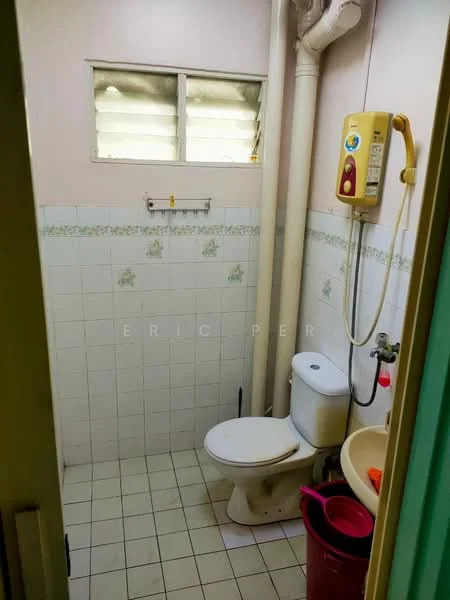 Bathroom features: white tiled walls, light grey tiled flooring, one-piece toilet, wall-mounted shower heater, handheld shower, sink with faucet, small window for ventilation, towel rack, plumbing pipes visible.