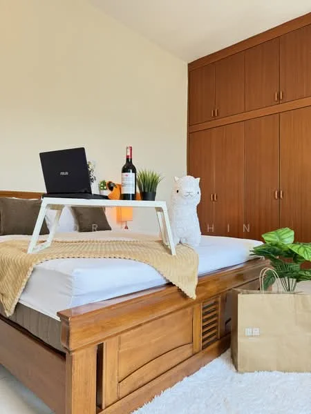 Bedroom features: queen-size bed, wooden bed frame, white bedding, decorative throw blanket, laptop on a tray table, wine bottle, stuffed animal, built-in wardrobe with wooden doors, green potted plant, paper bag on the floor, neutral wall color, natural light from window; style: modern.