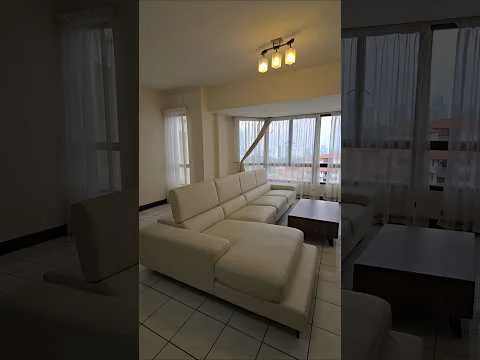 Living features: white leather sectional sofa, wood coffee table, floor-to-ceiling windows, sheer curtains, tile flooring, recessed ceiling lights, neutral painted walls, city view; style: modern; view: city.