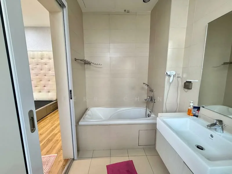 Bathroom features: bathtub, single-sink vanity with white cabinet, chrome basin mixer faucet, framed mirror, tile cladding on walls, cream tiled flooring, shower head, towel rack, soap dispenser, pink bath mat; style: modern. Room features: wood flooring, bed visible in background.