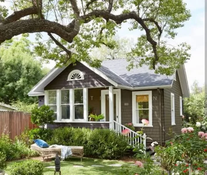 Exterior type: detached bungalow; features: wooden shingle facade in dark brown, white trim around windows and doors, gabled roof with dark shingles, arched window above the front door, front porch with white railing, manicured lawn, flower beds with blooming roses, mature trees providing shade, decorative outdoor furniture, garden pathway; style: traditional.