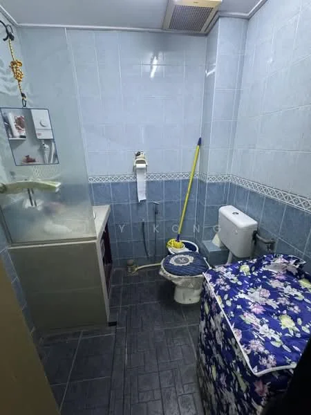 Bathroom features: blue tiled walls, grey tile flooring, single-sink vanity with cabinet, toilet, shower area with glass partition, mirror above vanity, toilet paper holder, cleaning supplies, decorative fabric covering on storage, wall-mounted faucet.