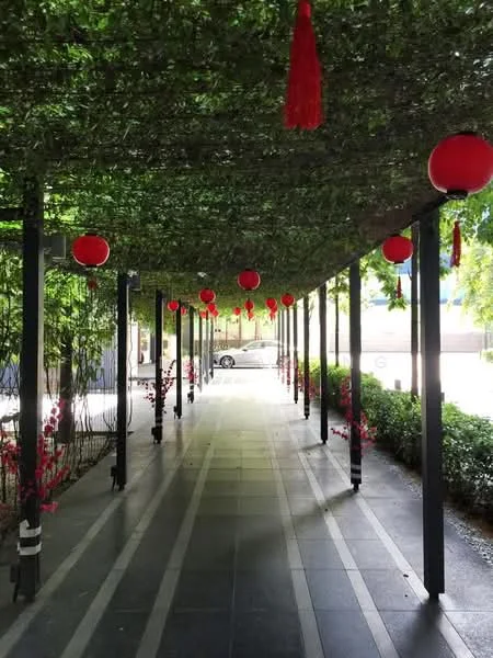 Exterior type: landscape garden; features: covered walkway with greenery, red lanterns hanging from the ceiling, decorative floral elements, paved stone flooring, surrounded by shrubs, ambient lighting, open-air design, columns supporting the structure; style: modern.