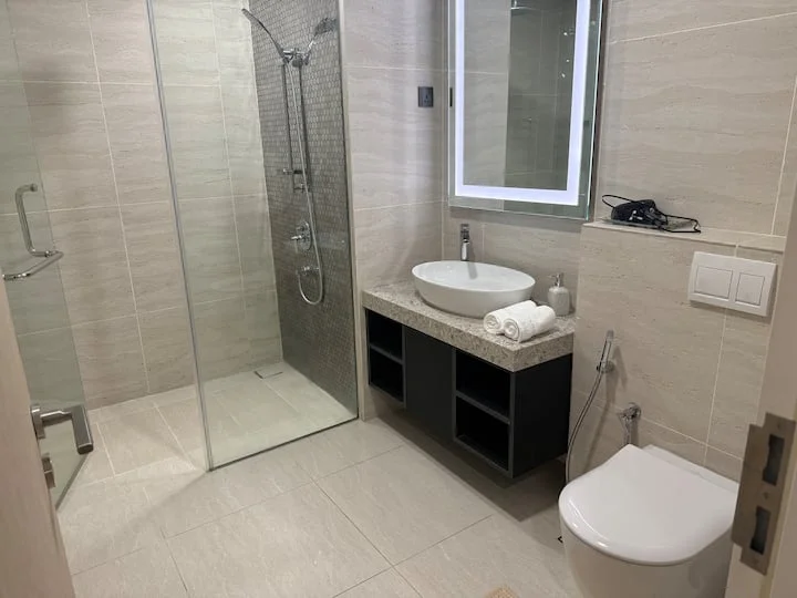 Bathroom features: walk-in shower with glass enclosure, rain shower head, handheld shower, round vessel sink, stone countertop, black cabinetry, wall-mounted mirror with LED lighting, one-piece toilet, towel rack, grey tile flooring, light beige wall tiles, shower wall with mosaic tile, chrome faucet; style: modern.