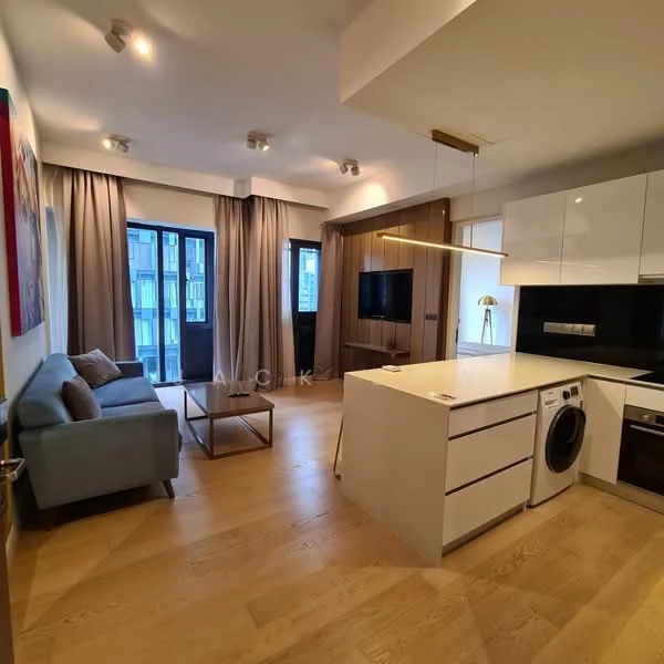 Living features: light wood flooring, white painted walls, floor-to-ceiling windows, sheer curtains, blue fabric sofa, coffee table, wall-mounted TV, recessed LED downlights, kitchen island with seating, built-in oven, stainless-steel washing machine, white cabinetry, black countertop; style: modern; view: city.