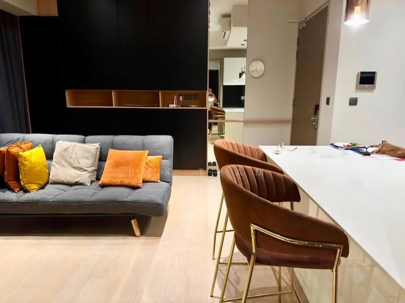 Living features: grey fabric sofa, decorative cushions in various colors, gold accent legs on sofa, white countertop, bar stools with brown upholstery and gold legs, wooden cabinetry, light wood flooring, wall-mounted clock, recessed lighting, dark feature wall, open layout to kitchen, neutral color palette; style: modern.