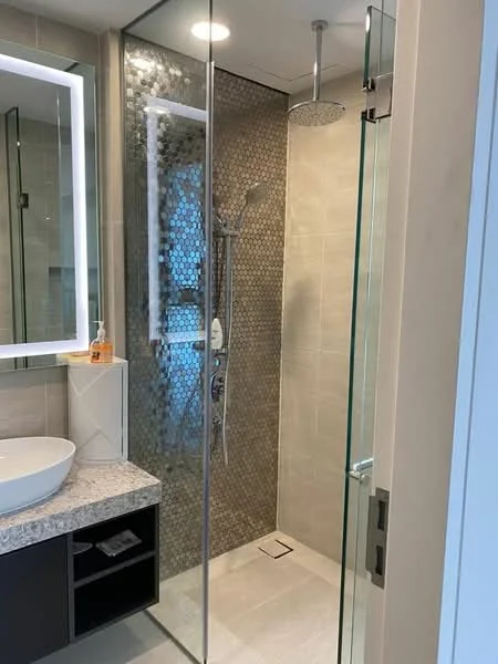Bathroom features: walk-in shower with glass enclosure, rain shower head, handheld shower, hexagonal tile wall cladding, light grey tile flooring, floating vanity with black cabinetry, white vessel sink, granite countertop, backlit mirror, recessed downlights; style: modern.