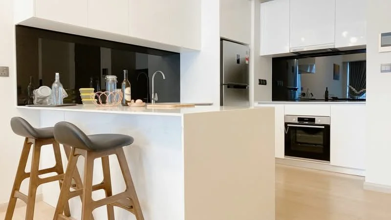 Kitchen features: white cabinetry with flat-panel doors, black glass backsplash, stainless-steel fridge, built-in oven, 4-burner gas hob, single-bowl stainless-steel sink, chrome pull-down faucet, countertop with breakfast bar, 2 wooden bar stools, recessed downlights, light wood flooring; style: modern.
