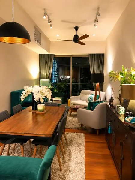 Dining features: wooden dining table, 6 upholstered dining chairs in teal, pendant lamp, recessed downlights, ceiling fan with 5 blades, area rug with neutral color, green decorative vase, white flowers in vase, sideboard with decorative items, floor-to-ceiling windows with curtains, green accent pillows on sofa, light-colored walls, wooden flooring; style: modern; view: city.