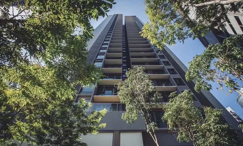 Exterior type: high-rise condominium; features: glass curtain wall, balconies on multiple floors, vertical fins, surrounded by greenery, tall trees in foreground, clear blue sky, modern architectural design; style: modern.