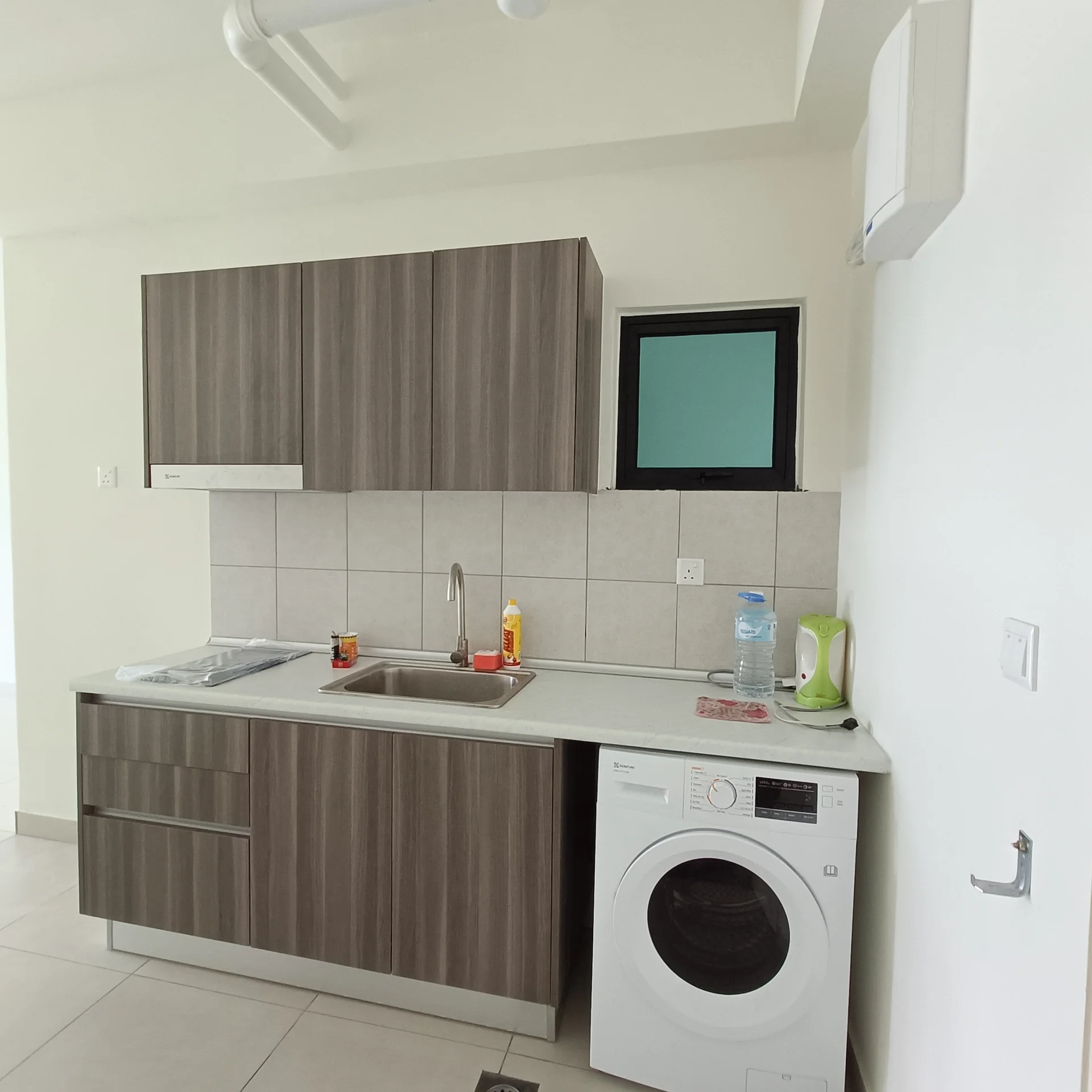 Kitchen features: brown wood cabinetry, white countertop, single-bowl stainless-steel sink, chrome pull-down faucet, built-in washing machine, tile backsplash, window above sink, kettle, dish soap, water dispenser; style: modern.