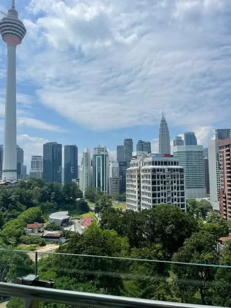 Exterior type: high-rise condominium; features: city skyline view, greenery in foreground, modern glass facades, Kuala Lumpur Tower visible, Petronas Twin Towers visible, various high-rise buildings, clear blue sky, lush landscaping; style: modern.