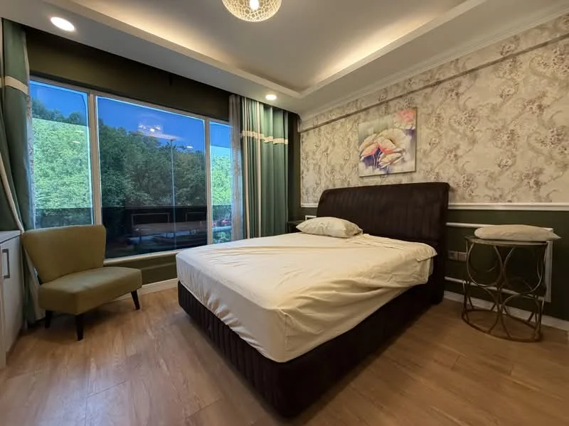 Bedroom features: queen-size bed with dark upholstered headboard, wooden flooring, green wallpaper with floral pattern, white painted walls with crown moulding, floor-to-ceiling windows, sheer curtains, decorative wall art, side table with metallic finish, armchair in green fabric, recessed ceiling lights, chandelier; style: contemporary; view: garden.