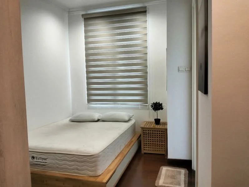 Bedroom features: queen-size bed with mattress, wooden bed frame, two white pillows, wooden bedside table, decorative plant in ceramic pot, brown wooden flooring, roller blinds on window, white painted walls, light fixture on ceiling; style: modern.