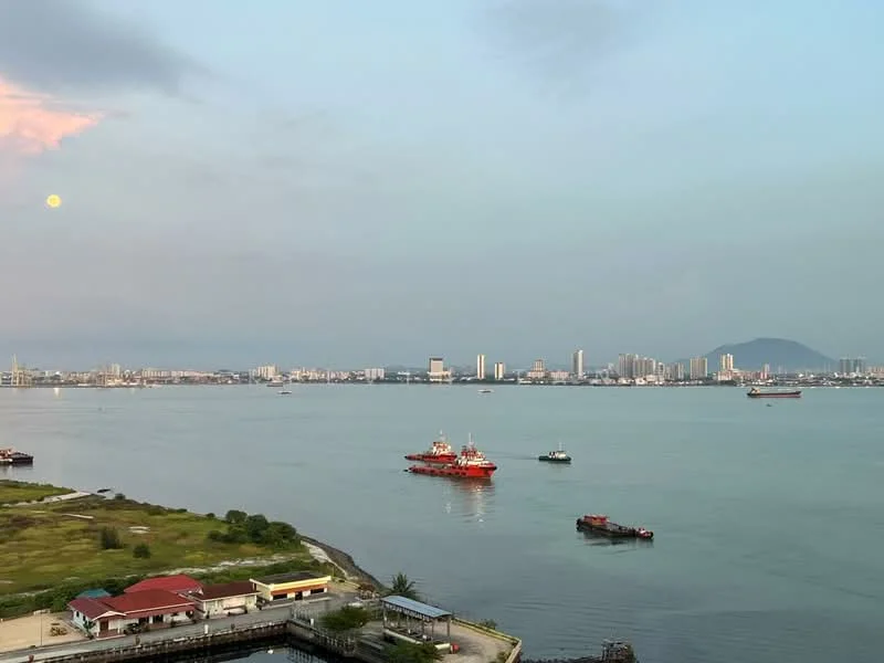 Exterior features: waterfront view, city skyline in background, boats on water, greenery along the shore, distant mountains, clear sky with clouds.
