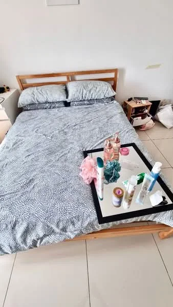 Bedroom features: queen-size bed with patterned bedding, wooden bed frame, two decorative pillows, nightstand with lamp, small bookshelf, various personal care products on tray, white walls, tile flooring; style: minimalist.