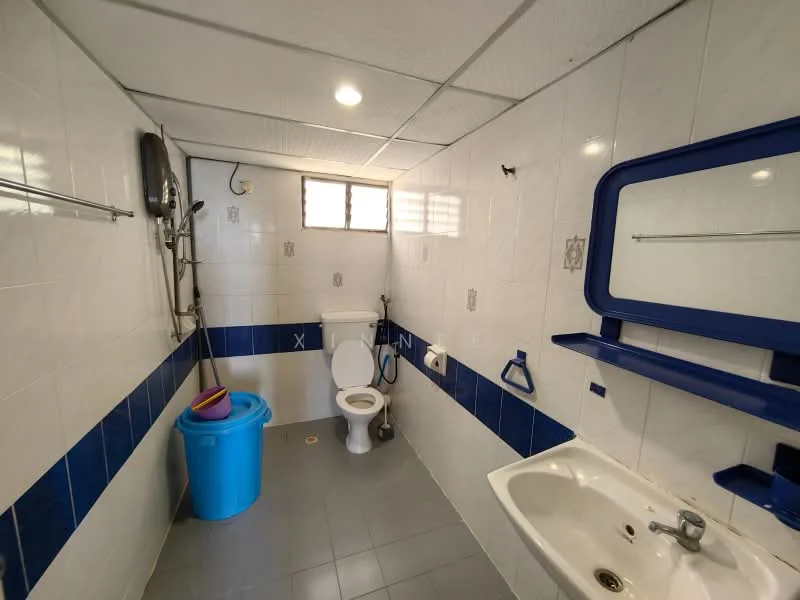 Bathroom features: white tiled walls, blue accent tiles, grey ceramic tile flooring, one-piece toilet, chrome basin mixer, white sink, mirror above sink, wall-mounted towel rack, shower area with handheld shower, window for natural light, blue plastic trash bin.