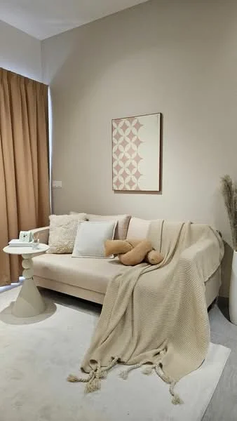 Living features: beige fabric sofa, decorative throw blanket, cream decorative pillows, tan curtains, round side table, framed wall art, plush teddy bear, light-colored area rug, neutral painted walls; style: modern.
