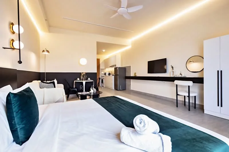 Room features: queen-size bed with green velvet throw, white bedding, 2 white towels rolled on bed, ceiling fan, recessed LED lighting, wall-mounted lamp, black accent wall, white wardrobe, wall-mounted TV, black countertop, stainless-steel fridge, kitchenette with built-in cabinets, dining table with white chair, decorative mirror; style: modern.