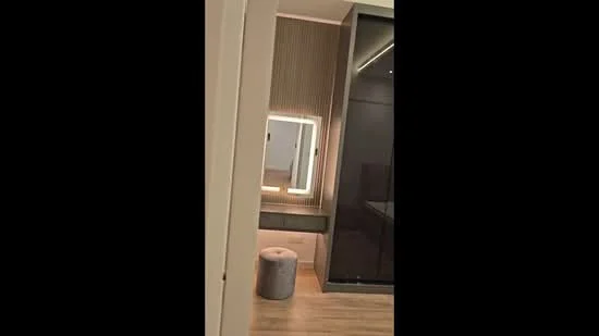 Room features: wooden flooring, backlit mirror, vanity table, stool, strip lighting, wall paneling, dark cabinetry, neutral wall color; style: modern.