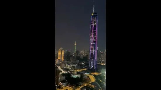 Exterior type: high-rise condominium; features: night view of city skyline, illuminated building facade, tall skyscraper with colorful lights, surrounding urban landscape, visible roads and traffic, adjacent buildings with varied heights; style: modern.