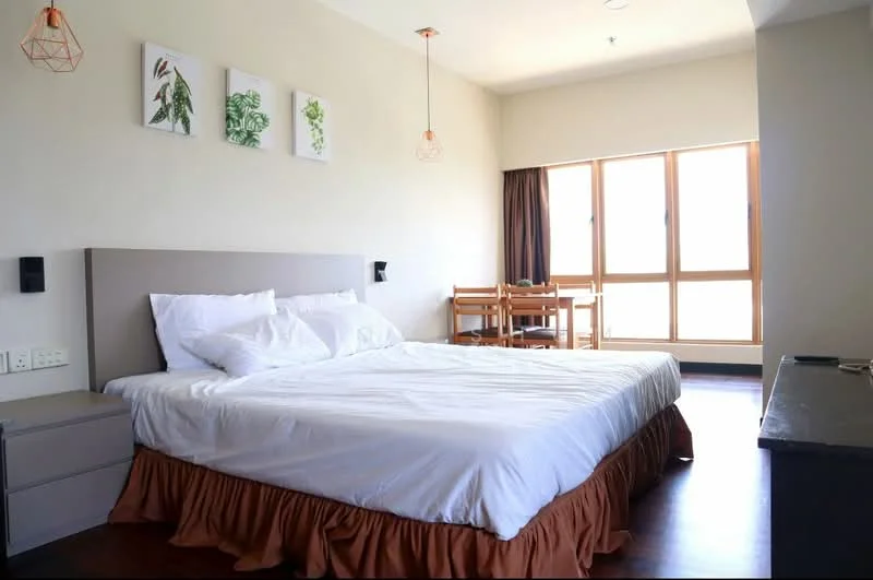 Bedroom features: king-size bed with white bedding, wooden bed frame, two bedside lamps, wooden bedside table, wall-mounted light fixtures, large windows with wooden frames, curtains in dark brown, decorative wall art with plants, dark wood flooring, dining table with chairs; style: modern.