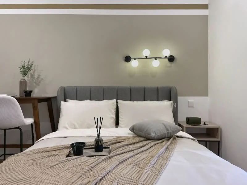 Bedroom features: queen-size bed, upholstered headboard in grey, white bedding with decorative throw, light beige knitted throw blanket, nightstand with small decorative items, table lamp with multiple bulbs, wall-mounted light fixture, neutral painted walls with accent stripes, wooden flooring, decorative reed diffuser, small potted plant; style: modern.