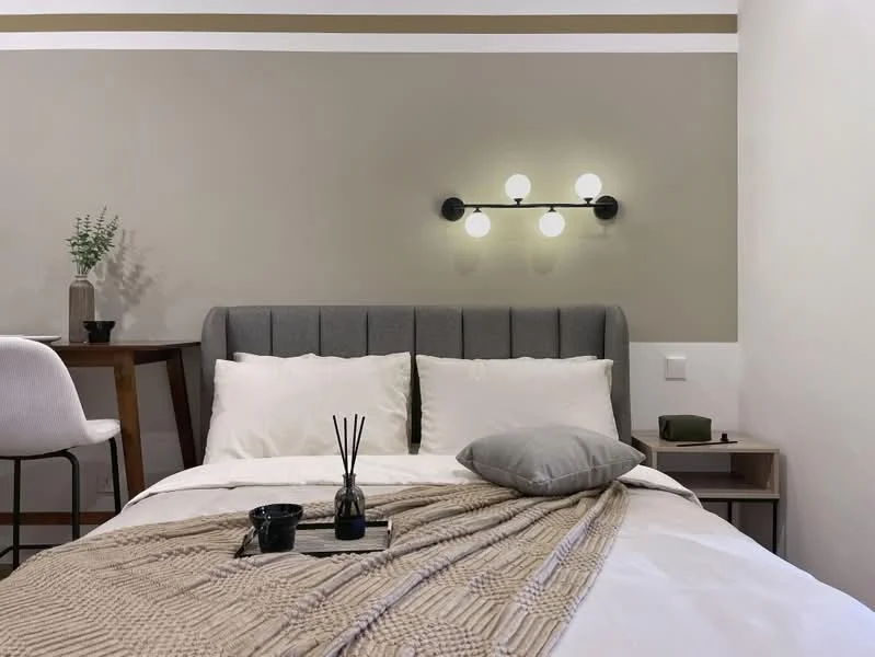 Bedroom features: queen-size bed, upholstered headboard in grey, white bedding with decorative throw, decorative throw pillow, nightstand with decorative items, table lamp, wall-mounted light fixture with multiple bulbs, neutral painted walls in beige and white, wooden flooring, small decorative plant, textured blanket; style: modern.