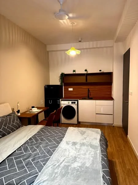 Room features: queen-size bed with grey geometric bedding, wooden bedside table, black refrigerator, white washing machine, white kitchen cabinetry, wooden countertop, black sink, single-bowl stainless-steel sink, chrome pull-down faucet, ceiling fan with light, wooden flooring, wall-mounted shelf with decorative plants, wall with vertical lines texture, light fixture with yellow shade; style: modern.