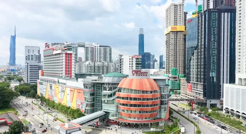 Exterior type: shopping mall; features: glass facade, orange and grey exterior cladding, circular dome structure, surrounded by high-rise buildings, multiple levels, large signage, urban context with busy road, green landscaping in front; style: modern.