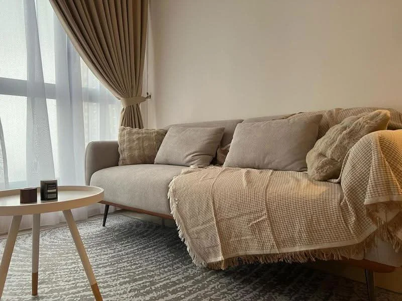 Living features: 3-seater fabric sofa in beige, decorative throw blanket, 2 decorative pillows, round coffee table with wooden legs, area rug with abstract pattern, floor-to-ceiling windows with sheer curtains, curtains in light brown, neutral wall color; style: modern.