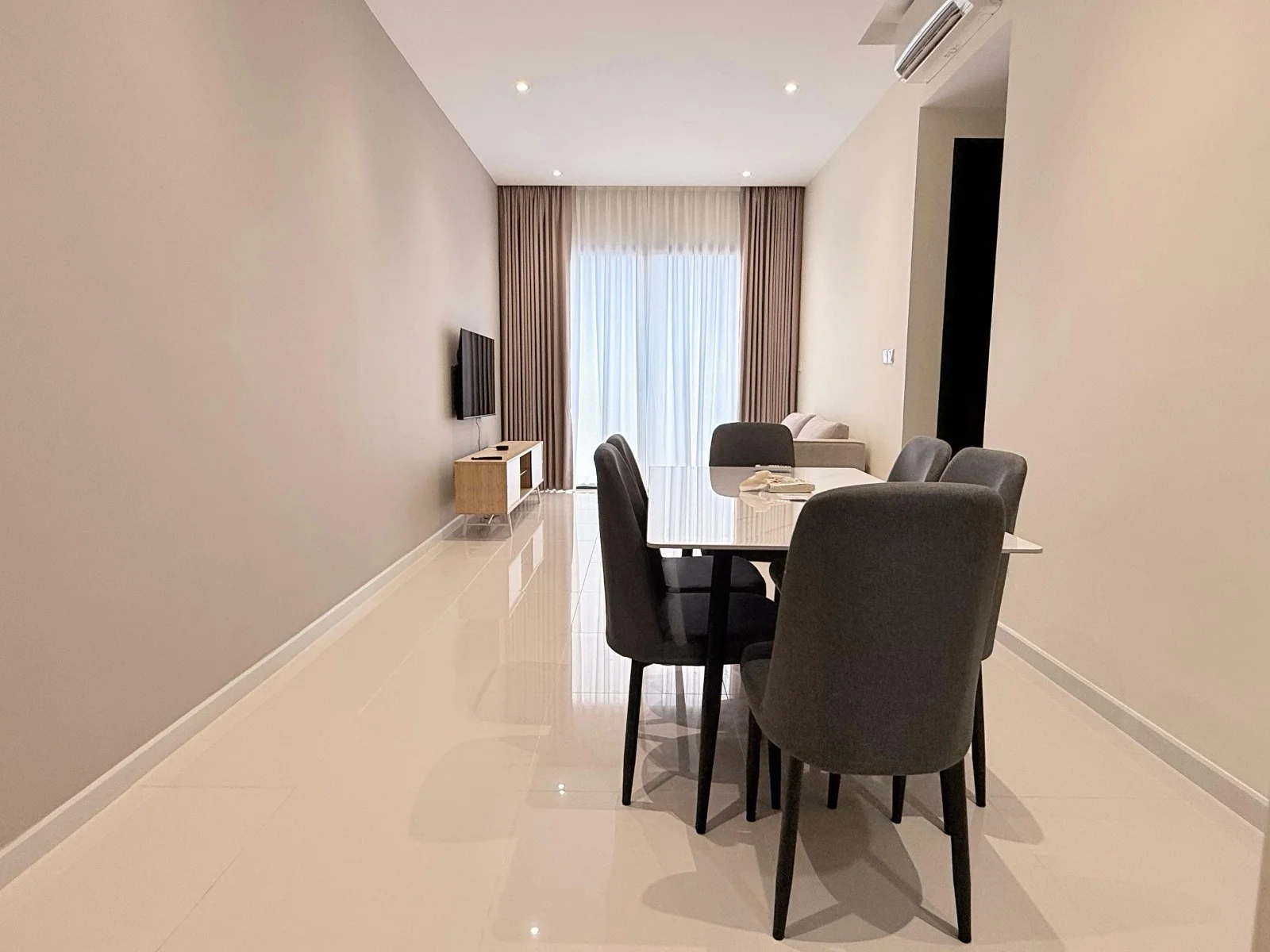 Dining features: white glossy tile flooring, light grey painted walls, recessed downlights, dining table with white top, 6 upholstered dining chairs in grey, wall-mounted TV, floor-to-ceiling windows with sheer curtains, curtains in light beige, minimalist wooden TV console; style: modern.