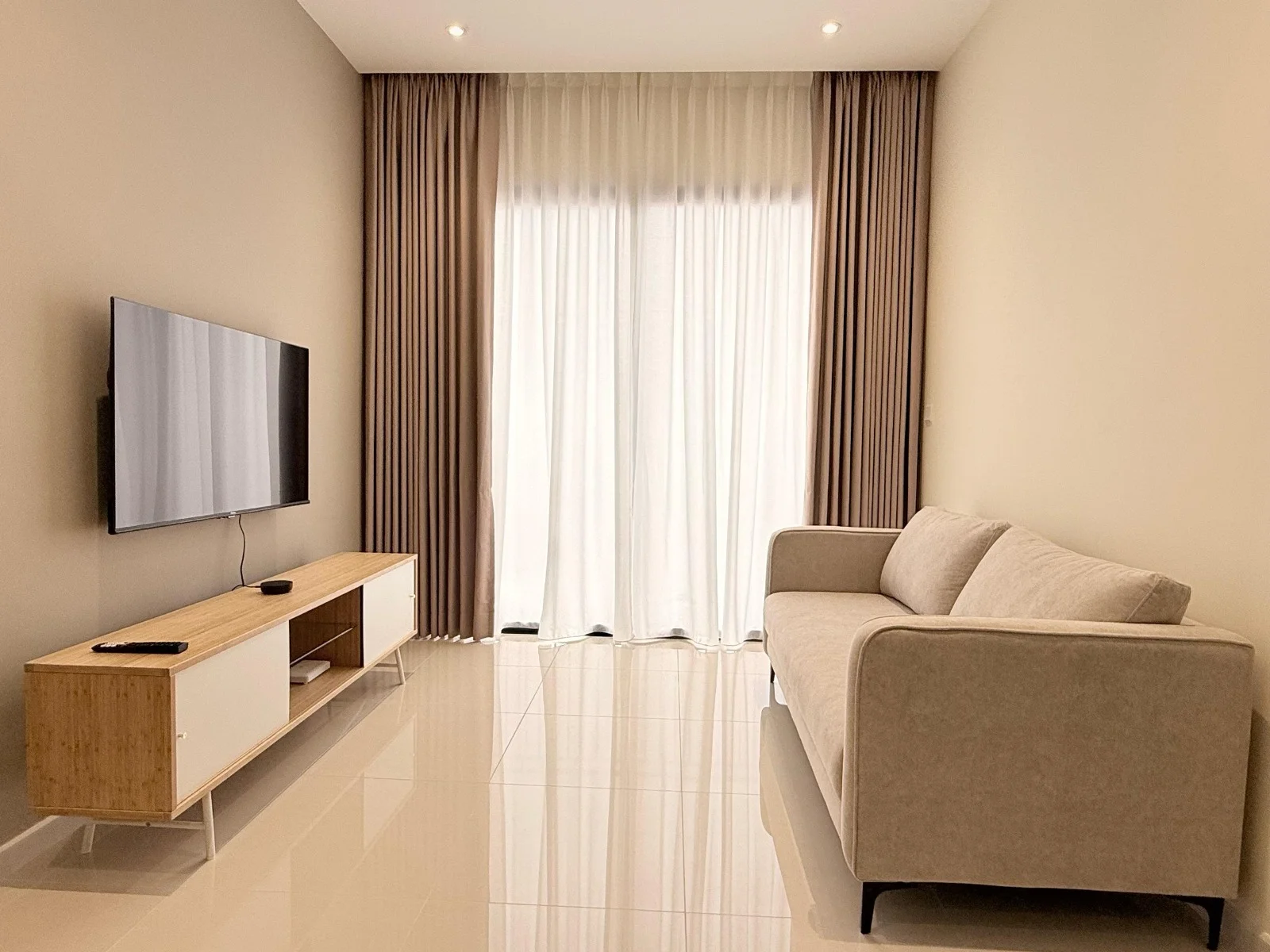 Living features: beige fabric sofa, wooden TV console with white cabinet, wall-mounted flat-screen TV, floor-to-ceiling windows, sheer curtains, cream painted walls, glossy tile flooring, recessed downlights; style: modern.