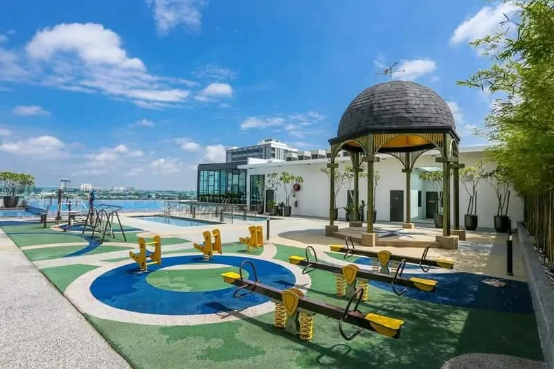 Exterior type: landscape garden; features: outdoor playground with equipment, decorative gazebo with dome roof, colorful rubberized flooring, manicured landscaping with potted plants, swimming pool in background, city skyline view, sunny blue sky, open space for activities; style: modern.