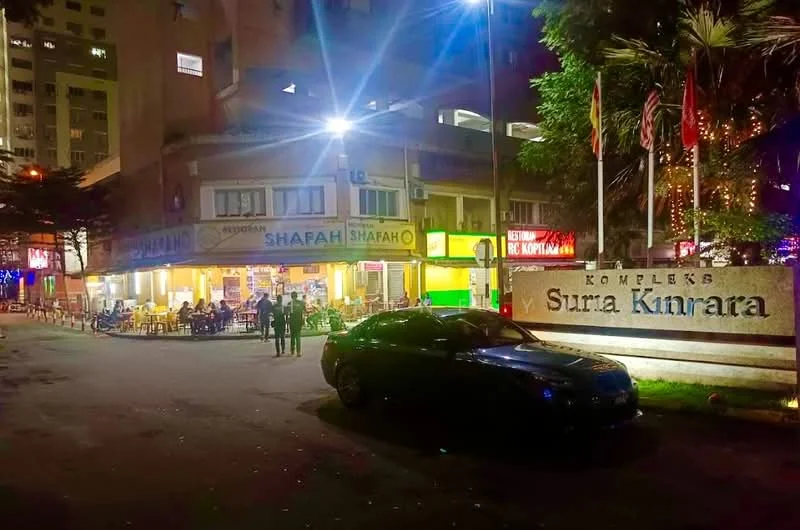 Exterior type: low-rise apartment block; features: illuminated signage, outdoor dining area, paved walkway, landscaped greenery, parking space, street lighting, multiple storefronts, entrance sign, adjacent to residential buildings; style: modern.