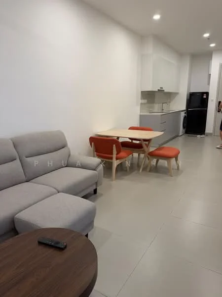 Living features: grey fabric sectional sofa, wood coffee table, light grey walls, recessed downlights, tile flooring, dining table with wooden legs, 2 upholstered dining chairs in orange, kitchen area visible in background, black refrigerator, washing machine, open layout; style: modern.