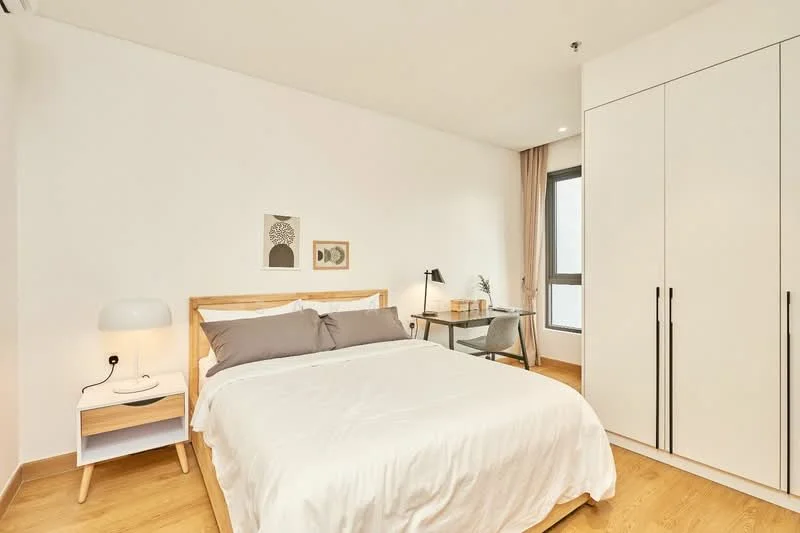 Bedroom features: queen-size bed with white bedding, wooden bed frame, white painted walls, light wood flooring, built-in wardrobe with white doors, desk with chair, table lamp, framed artwork on wall, window with sheer curtains; style: modern.