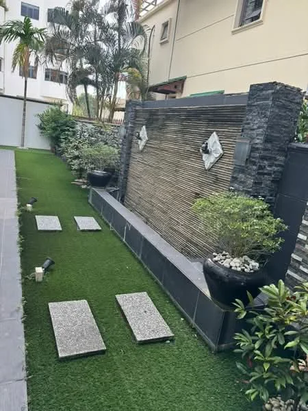 Exterior type: landscape garden; features: artificial grass lawn, stone pathway, water feature with stone cladding, decorative wall with diamond-shaped accents, potted plants, landscaped shrubs, outdoor lighting fixtures; style: modern.