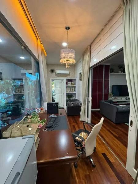 Study features: wooden flooring, chandelier light fixture, wall-mounted air conditioning unit, large window with sheer curtains, desk with computer setup, white office chair, decorative shelving units, wall decor with framed photos, open doorway leading to living area; style: modern; view: garden.