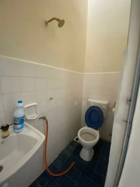 Bathroom features: white tiled walls, blue floor tiles, toilet, sink, shower head, exposed plumbing, cleaning supplies, hose, mirror, wall-mounted soap dispenser.