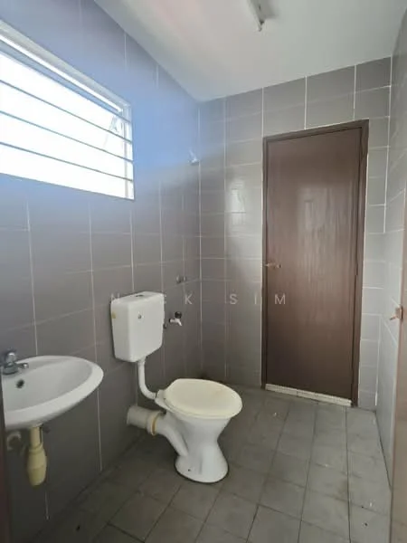 Bathroom features: grey tiled walls, white ceramic toilet, white pedestal sink, single-lever faucet, window with horizontal slats, light fixture on ceiling, grey tiled flooring, wooden door.