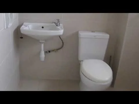 Bathroom features: white ceramic toilet, wall-mounted sink, chrome faucet, light beige tiled walls, light beige tiled flooring, exposed plumbing under sink.