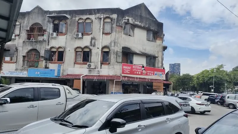 Exterior type: shophouse; features: weathered facade, multiple air conditioning units, red signage, ground floor retail space, first floor residential units, balconies with wrought iron railings, white painted walls, surrounding parking area, urban context with nearby buildings; style: traditional.