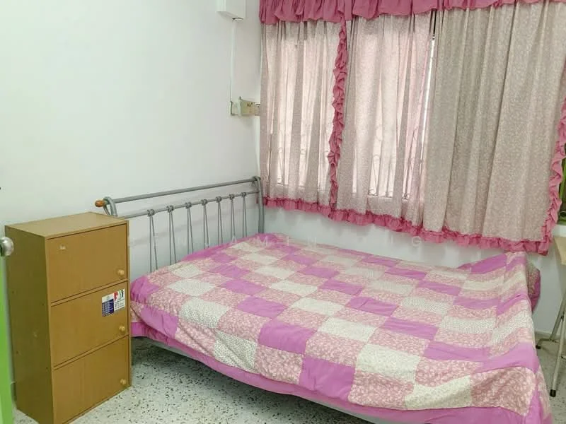 Bedroom features: metal bed frame with mattress, pink and white checkered bedspread, curtains with ruffled edges in pink, single bedside cabinet with drawers, textured flooring, wall-mounted electrical outlet, window with natural light, decorative wall with minimal decor.