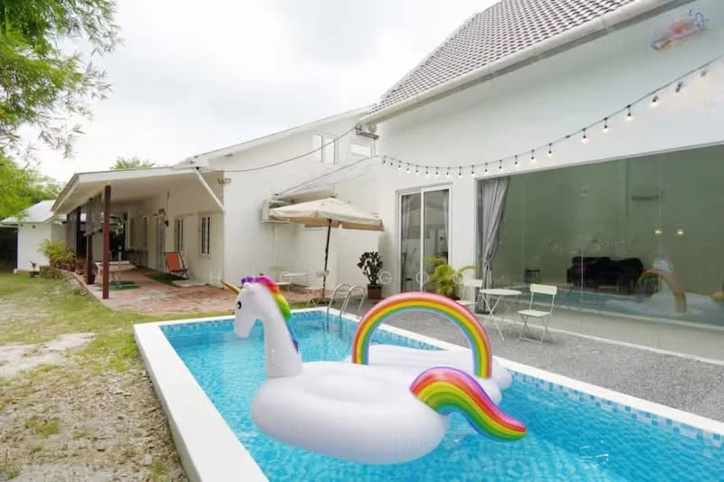 Exterior type: detached bungalow; features: white painted facade, flat roof, swimming pool with inflatable unicorn, outdoor dining area with chairs, umbrella for shade, string lights for ambiance, landscaped garden with gravel pathway, patio area with seating, large glass windows, manicured lawn; style: modern.