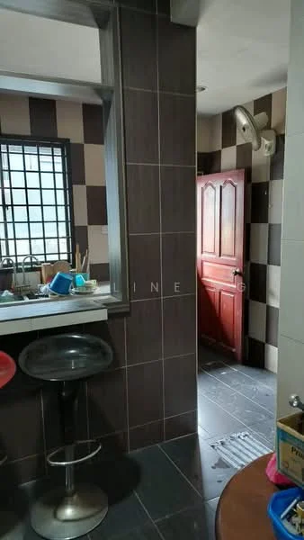 Kitchen features: black and brown tiled walls, grey tiled flooring, bar counter with two stools, sink, window with grid pattern, kitchen utensils on counter, door leading to another room, exhaust fan. Room features: wooden door, grey tiled flooring, brown tiled walls.